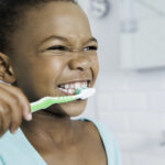 Young Black girl smiles as she brushes her teeth with a green toothbrush in the bathroom