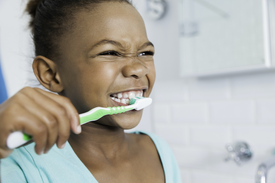Young Black girl smiles as she brushes her teeth with a green toothbrush in the bathroom