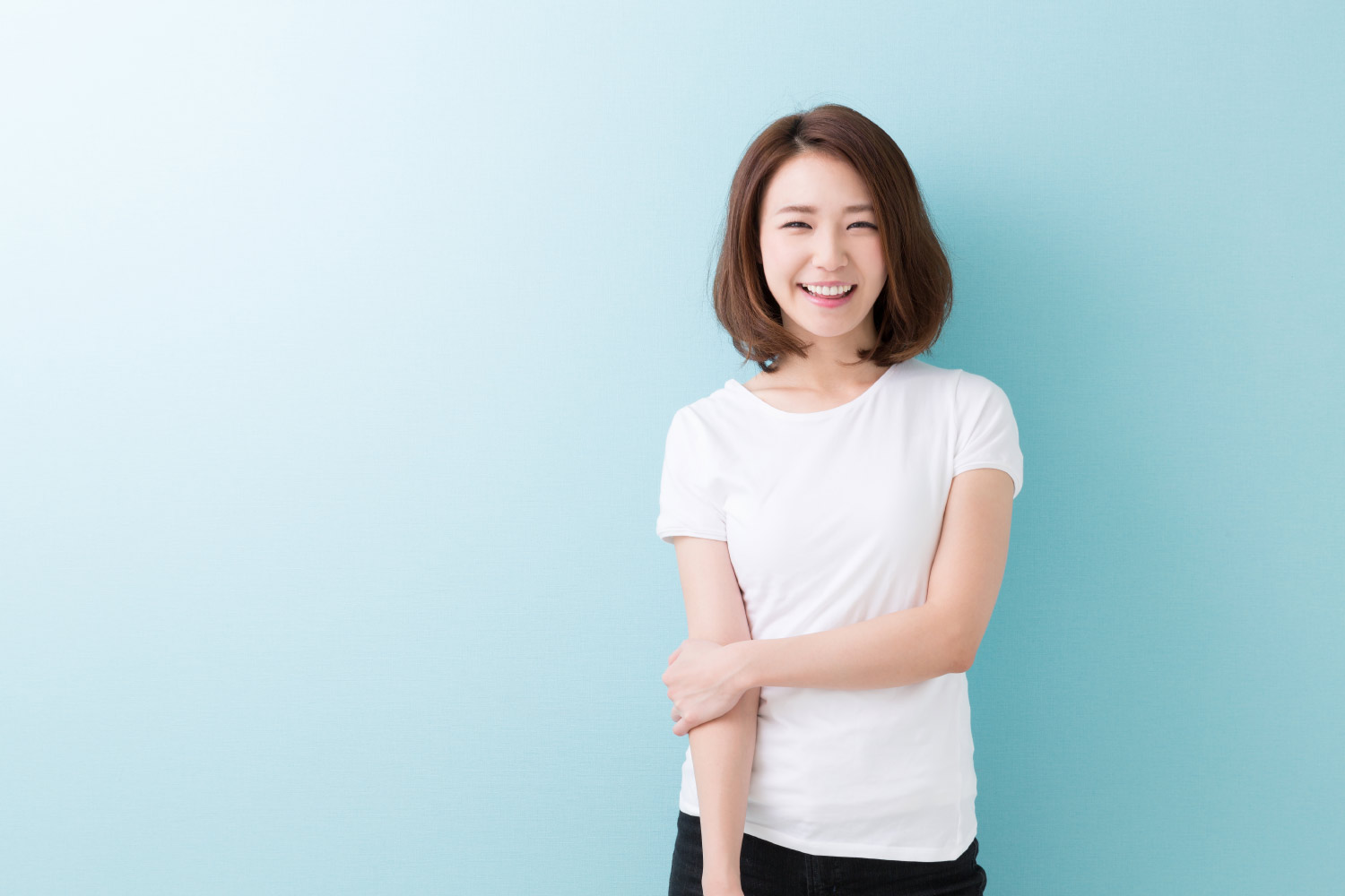 Asian woman in a white t-shirt smiles against a blue wall after professional teeth whitening